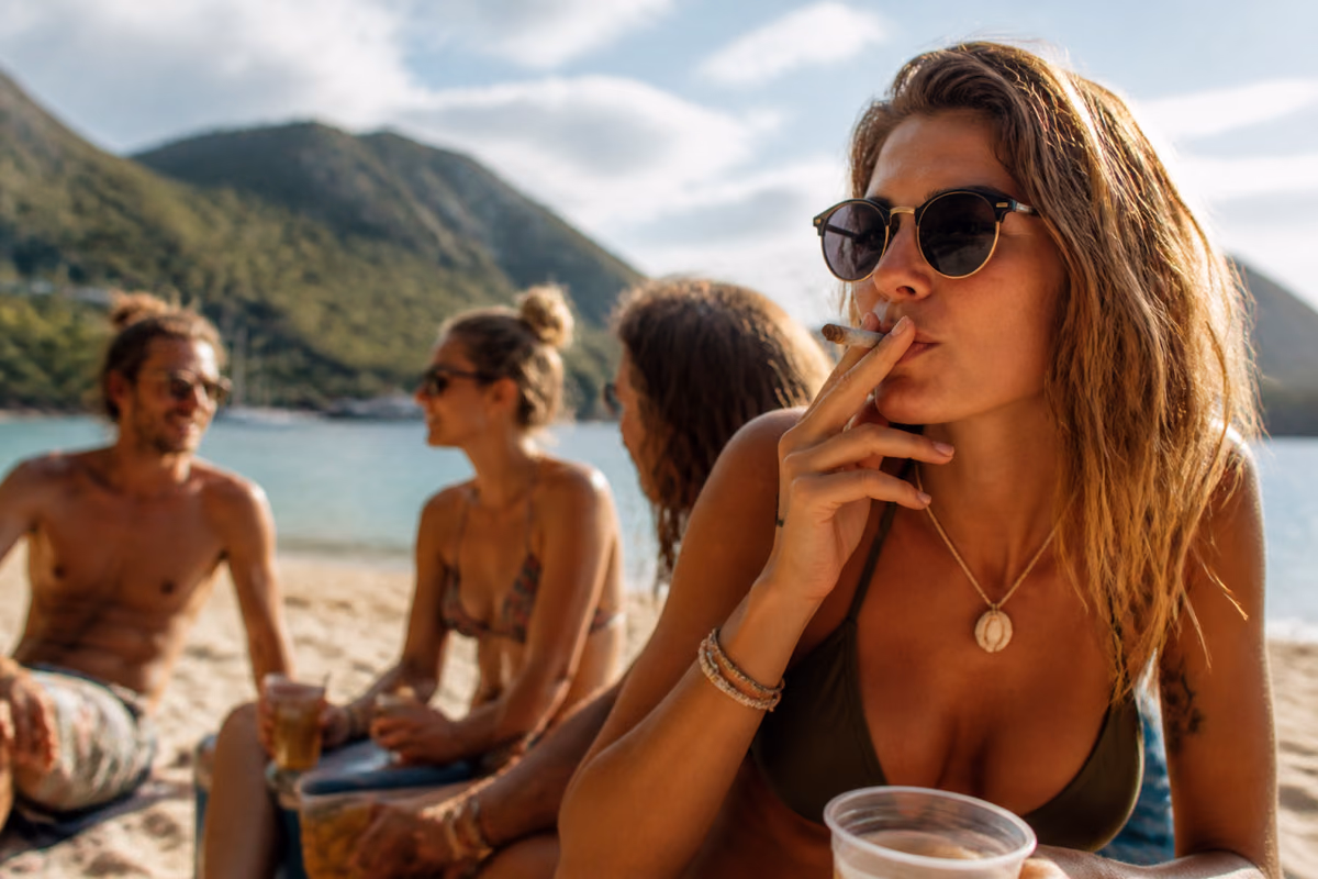Žena so slnečnými okuliarmi fajčí joint na tropickej pláži, v pozadí priatelia s nápojmi
