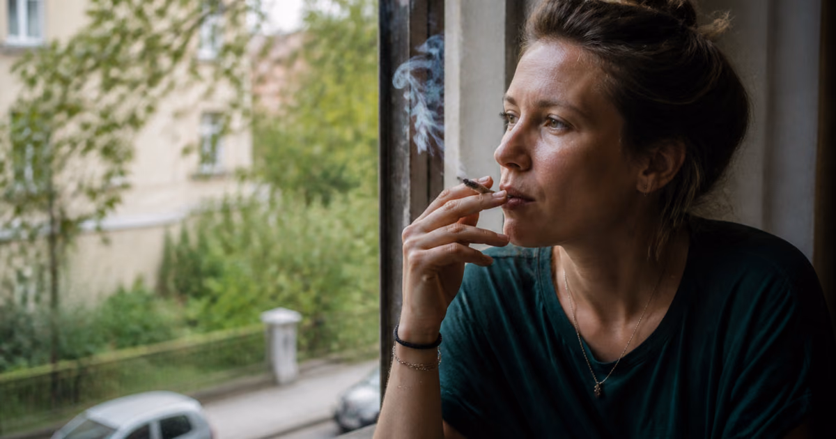 Žena fajčí joint pri okne a pozerá sa na ulicu plnú zelene